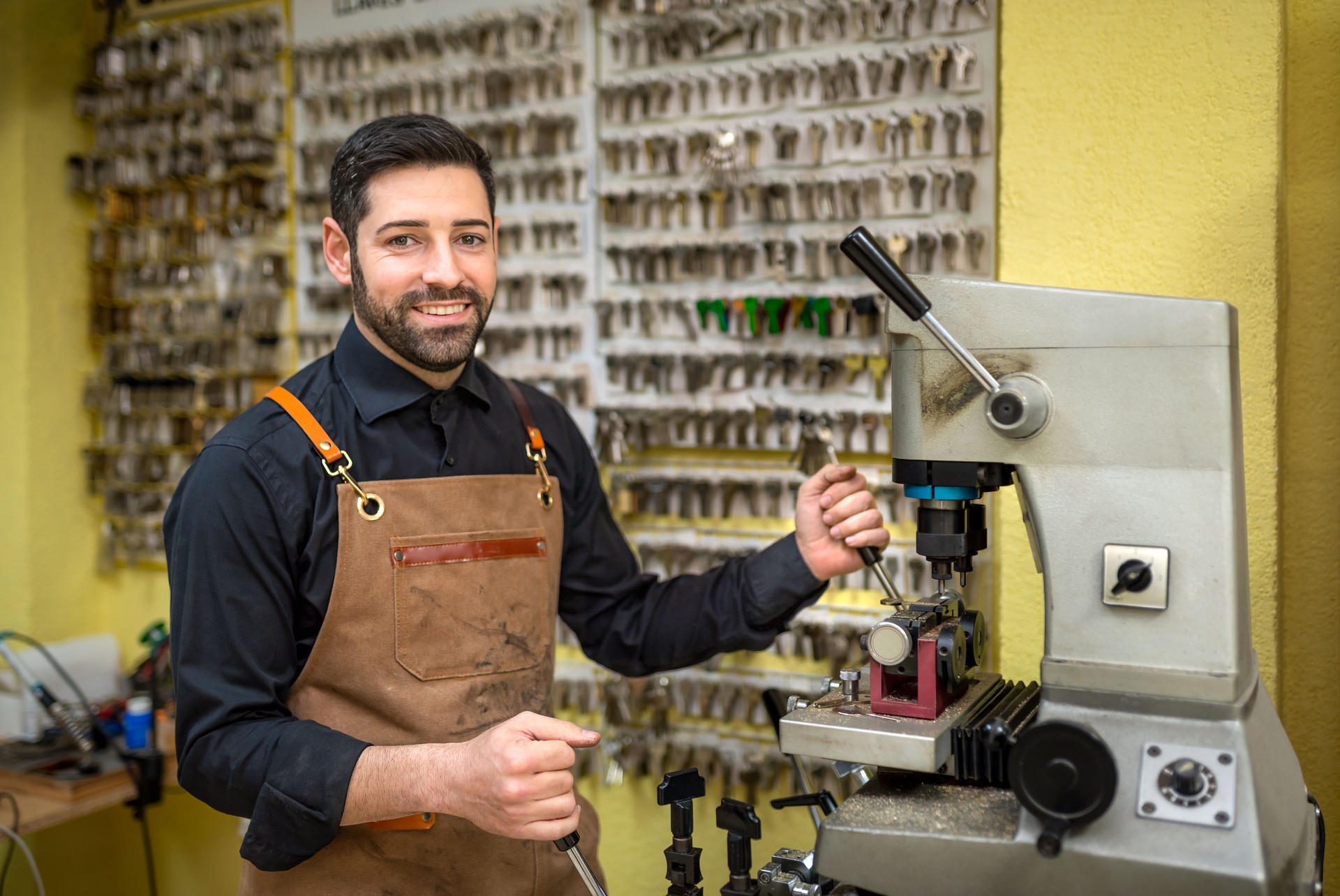Locksmith smiling while duplicating metal keys on copying machine, wearing protective apron in professional workshop setting